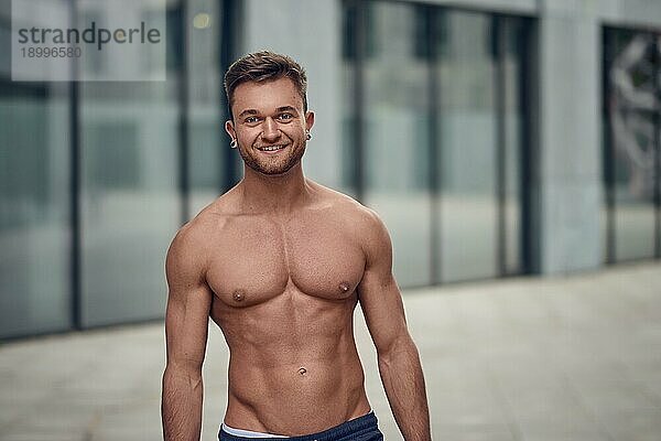 Attraktiver  muskulöser junger Mann  der ohne Hemd in einer städtischen Umgebung posiert und mit einem warmen  freundlichen Lächeln in die Kamera schaut  mit Kopierraum