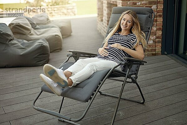 Glücklich lächelnde attraktive junge schwangere junge Frau  die sich im Freien auf einem Liegestuhl auf einer Holzterrasse entspannt und ihren Babybauch in den Händen wiegt