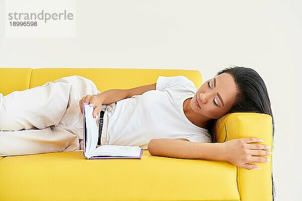 Hübsche asiatische Frau  die auf einer gemütlichen gelben Couch liegend ein Buch liest  Konzepte von Heim und Komfort  Hobby  Freizeit