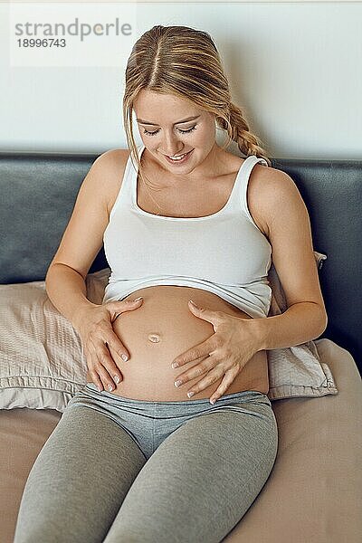 Glücklich lächelnde attraktive junge schwangere Frau  die ihren nackten Babybauch in den Händen wiegt  während sie sich auf einem Bett entspannt und in die Kamera schaut
