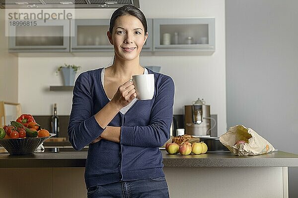 Glückliche  zufriedene Hausfrau in ihrer Küche  die der Kamera ein warmes  freundliches Lächeln schenkt  während sie sich mit einer Tasse Kaffee und frischem Obst auf dem Tresen hinter ihr entspannt