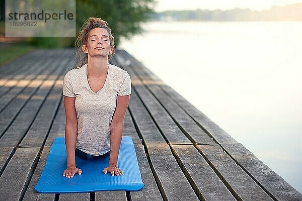 Attraktive junge Frau  die auf einer Matte auf einem hölzernen Deck über einem See oder Fluss trainiert und eine Yogapose mit geschlossenen Augen und heiterem Gesichtsausdruck einnimmt