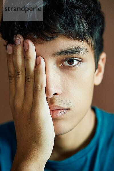 Close up Porträt eines jungen Mannes  der sein halbes Gesicht mit einer Hand bedeckt und mit einem Auge auf einen Studiohintergrund schaut. Mann Schönheit Konzept