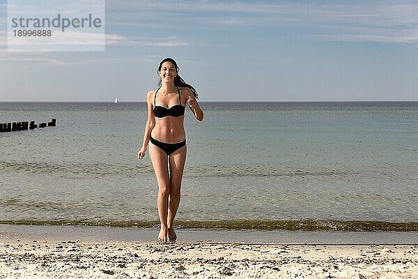 Lachende schöne junge Frau in einem schwarzen Bikini  die vor Freude springt und ihr langes brünettes Haar in die Luft wirft  an einem tropischen Strand mit einem ruhigen Ozean im Hintergrund mit Kopierraum