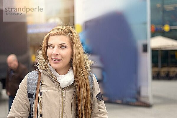 Freundliche attraktive blonde Frau mit Brille in warmer Wintermode steht in einer städtischen Straße mit hellen Sonnenstrahlen über ihrem Kopf