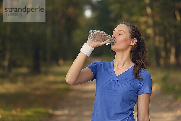 Fitte schlanke junge Frau in Sportkleidung trinkt Wasser in Flaschen  während sie auf einem Waldweg eine Pause einlegt  um während eines Trainingslaufs auf dem Lande neue Flüssigkeit zu sich zu nehmen  Gesundheits und Fitnesskonzept