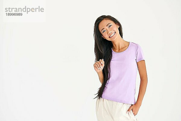Hübsche lächelnde asiatische Frau in lässiger Kleidung posiert mit Blick in die Kamera auf weißem Hintergrund mit Kopie Raum