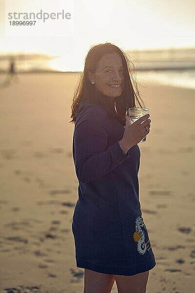 Frau genießt den Sonnenuntergang an einem tropischen Strand  während sie einen Cocktail trinkt und sich umdreht  um mit einem fröhlichen  lebhaften Lächeln über ihre Schulter in die Kamera zu schauen