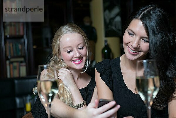 Zwei schöne junge Frauen genießen ein Glas Wein in einer Nacht mit ihren Handys und lachen über das Gespräch und die Textnachricht
