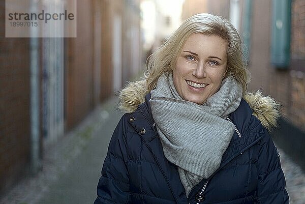 Attraktive  freundliche blonde Frau mit modischem Winterschal  die in einer engen Gasse in der Stadt steht und fröhlich in die Kamera lächelt