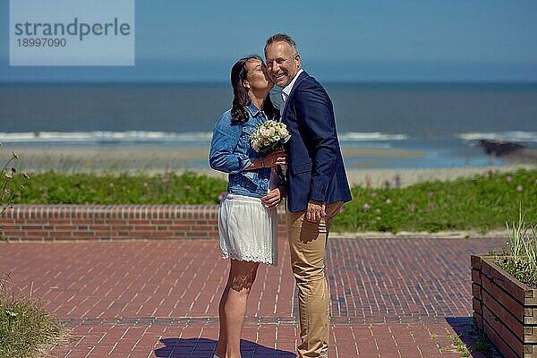 Glücklich lächelndes Hochzeitspaar  das in die Kamera schaut  während sie Hand in Hand auf einem Steg stehen und einen Blick auf einen tropischen Strand und das Meer während ihres romantischen Ausflugs genießen
