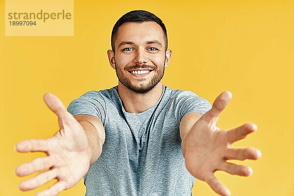 Hübscher glücklicher Mann mit offener Hand bereit für Umarmungen auf gelbem Hintergrund. Emotionen Konzept