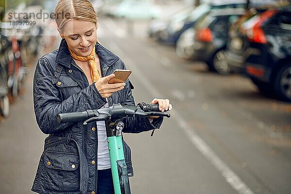 Glückliche junge Frau in lässiger grauer Jacke  die in die Kamera schaut und lächelt  steht auf der Straße mit einem escooter und einem Handy in der Hand