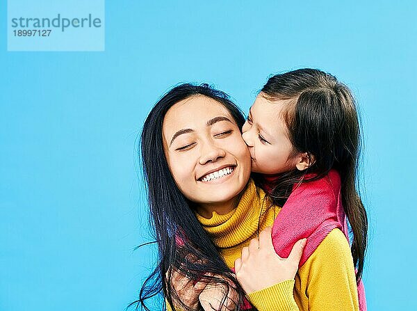 Kleine süße Tochter umarmen und küssen ihre glückliche Mutter vorblauem Hintergrund. Familie Liebe Konzept