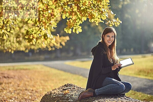 Hübsche junge Frau entspannt sich in einem herbstlichen Park  liest auf ihrem Tablet PC und lächelt fröhlich in die Kamera