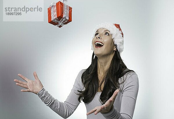 Lachende schöne Frau mit roter Weihnachtsmannmütze  die mit freudiger Erregung ein in der Luft schwebendes Weihnachtsgeschenk über ihren ausgestreckten Händen auffängt