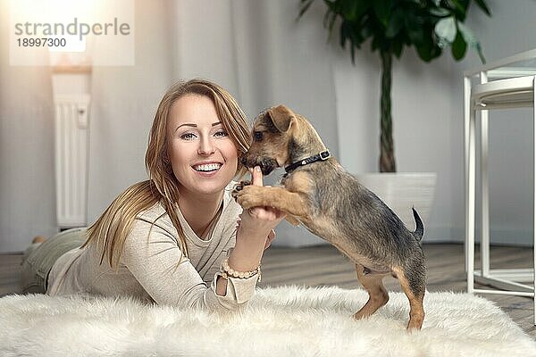 Attraktive freundliche Frau  die mit ihrem kleinen Hund auf einem flauschigen Teppich auf einem Holzboden im Wohnzimmer kuschelt und dabei glücklich in die Kamera lächelt