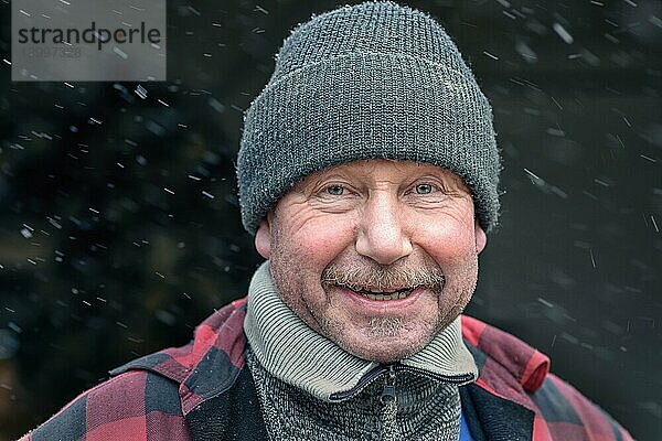 Glücklicher Mann mit Schnurrbart in einer gestrickten Wintermütze und warmer Winterkleidung  der mit einem charismatischen  freundlichen Lächeln in die Kamera blickt  während er im Freien im Schnee steht