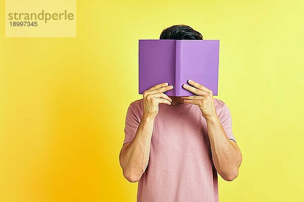 Junger Mann versteckt sein Gesicht hinter einem Buch auf gelbem Hintergrund. Emotionen Konzept