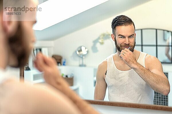 Attraktiver junger bärtiger Mann  der sich im Badezimmer die Zähne putzt und sich dabei im Spiegel betrachtet  in einem Zahnpflegekonzept