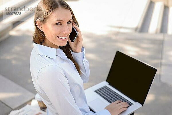 Blick von oben auf eine freundliche  attraktive junge Frau  die in einem Kommunikations und Mobilitätskonzept im Freien mit Kopierraum an einem Laptop arbeitet und mit einem Mobiltelefon spricht