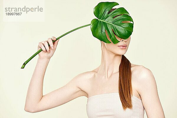 Schöne junge Frau bedeckt und versteckt Gesicht mit monstera tropischen Blatt. Naturkosmetik  Hautpflege  Spa  gesichtslose Person Konzept