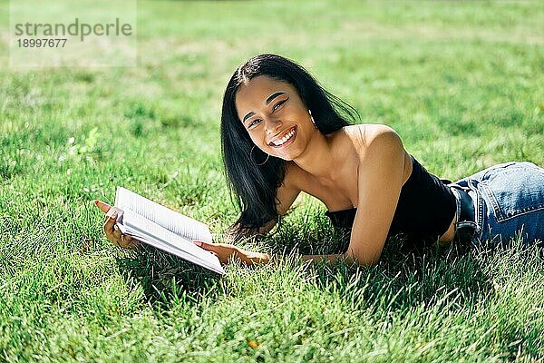 Hübsche afroamerikanische Frau liest Buch im Gras liegend im Park. Entspannen und Hobby Konzept