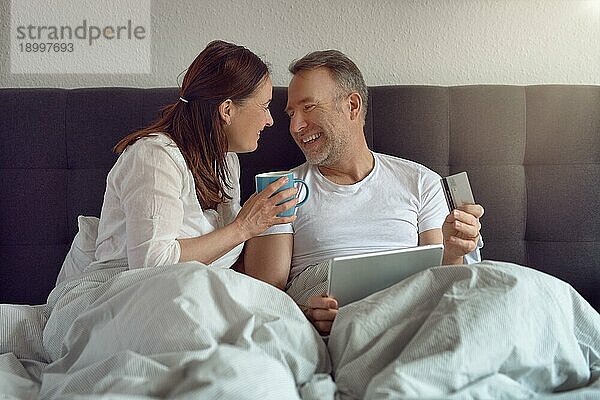 Glückliches Paar mittleren Alters  das morgens im Bett sitzt  Kaffee trinkt und mit einem digitalen Tablet online einkauft