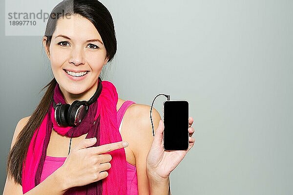 Porträt einer attraktiven  fröhlichen brünetten jungen Frau  die Kopfhörer um den Hals trägt und auf ihr intelligentes Mobiltelefon zeigt  auf grauem Hintergrund