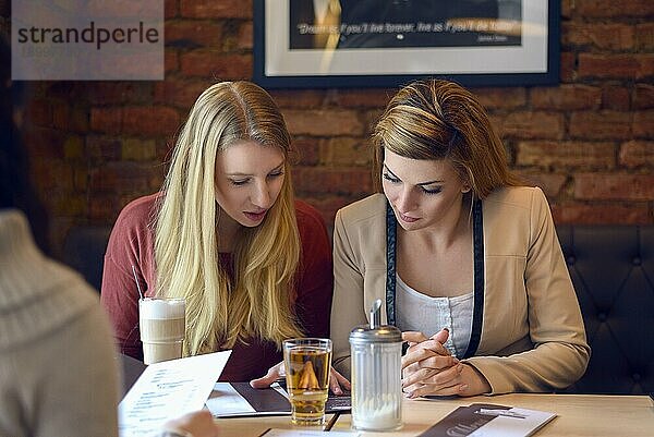 Zwei schöne blonde junge erwachsene Frauen  die an einem Stand sitzen und ihre Speisekarte in einem Restaurant mit einer Backsteinmauer hinter ihnen durchgehen
