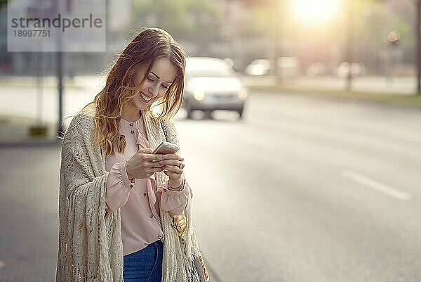 Attraktive junge Frau  die mit einem glücklichen Lächeln eine Nachricht auf ihrem Mobiltelefon überprüft  während sie am Rande einer städtischen Straße im frühen Morgenlicht mit Kopierraum steht