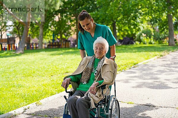 Eine ältere Frau mit der Krankenschwester auf einem Spaziergang durch den Garten eines Pflegeheims in einem Rollstuhl neben der Natur und den Bäumen