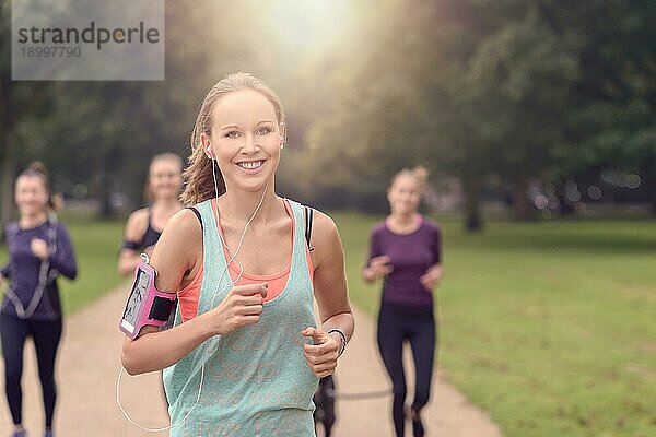 Halbkörperaufnahme einer sportlichen  hübschen jungen Frau  die in die Kamera lächelt  während sie mit anderen Mädchen im Park joggt  mit Kopierbereich auf der rechten Seite