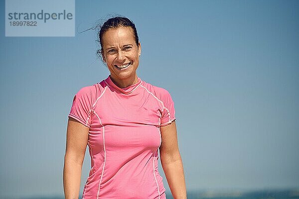 Glücklich lächelnde attraktive sportliche schlanke Frau mittleren Alters  die mit einem warmen freundlichen Lächeln in die Kamera blickt  vor einem sonnigen blaün Sommerhimmel mit Kopierraum