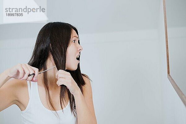 Attraktive junge Frau mit langen brünetten Haaren  die sich darauf vorbereitet  sie mit einer Schere im Badezimmer abzuschneiden  und dabei ängstliche Grimassen schneidet  da sie Zweifel an ihrer Entscheidung hat