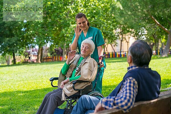 Eine ältere Frau mit der Krankenschwester bei einem Spaziergang durch den Garten eines Pflegeheims im Rollstuhl und beim Begrüßen eines älteren Mannes