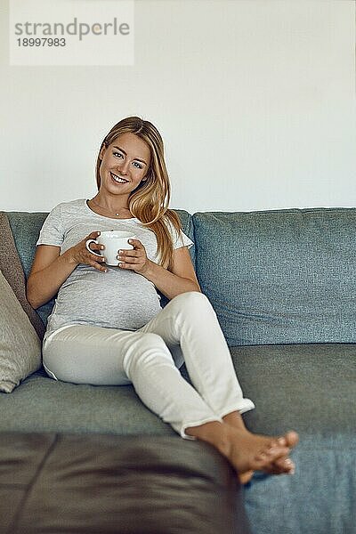 Hübsche barfuß junge schwangere Frau  die sich auf einer Couch entspannt  eine Tasse heißen Tee trinkt und glücklich in die Kamera lächelt
