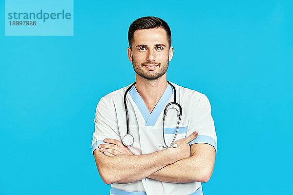 Porträt eines zuversichtlich lächelnden männlichen Arztes mit Stethoskop in medizinischer Uniform auf blauem Hintergrund mit verschränkten Armen