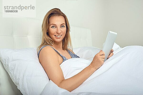 Attraktive Frau entspannt sich in einem bequemen neutralen weißen Bett mit einem Tabletcomputer in den Händen und einem freundlichen Lächeln