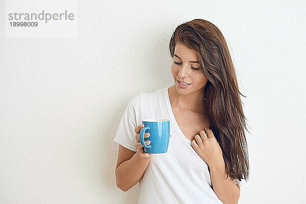 Junge brünette Frau in weißem TShirt die mit beiden Händen aus einem blaün Becher trinkt und mit einem freundlichen Lächeln in die Kamera schaut. Stehend gegen weiße Wand mit Kopie Raum