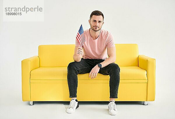 Junge fröhliche Mann mit amerikanischer Flagge in der Hand sitzt auf hellen Sofa decken weißem Hintergrund. Independence Day  Flag Day Konzept