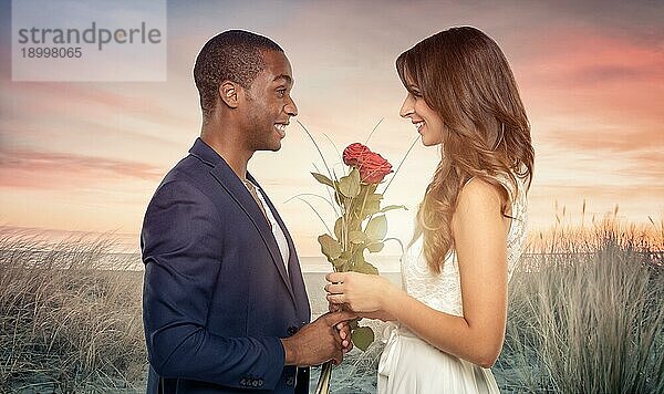 Lächelnder  romantischer  gutaussehender junger afrikanischer Mann macht seiner Liebsten einen Heiratsantrag  während er einen Strauß langstieliger roter Rosen in der Hand hält  mit Blick auf den Sonnenuntergang am Strand