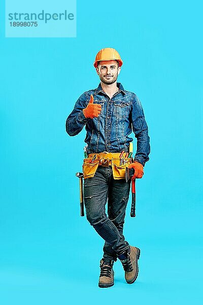 Porträt eines selbstbewussten  gut aussehenden männlichen Bauarbeiters mit Schutzhelm  der ein Daumen hoch Zeichen vor blauem Hintergrund zeigt und in die Kamera blickt  in voller Länge