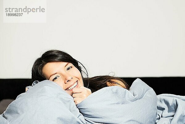 Lachende attraktive junge Frau  die sich in ihrem gemütlichen warmen Bett einkuschelt und über die Bettdecke hinweg in die Kamera blickt