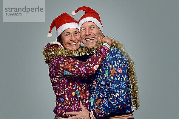 Ein fröhliches Ehepaar feiert Weihnachten zusammen in festlichen roten Weihnachtsmannmützen und Weihnachtsmänteln  die sich umarmen und lachen  während sie in die Kamera lächeln
