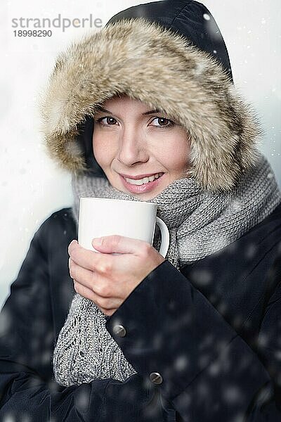 Hübsche junge Frau in warmer Wintermode  warm eingepackt in einen dicken Mantel  einen Strickschal und eine Pelzmütze  die mit einem genussvollen Lächeln einen Becher mit heißer Suppe oder Kaffee in der Hand hält