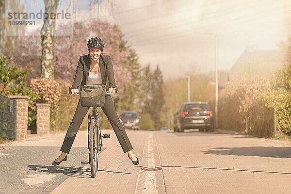 Elegante Frau in einem lockeren Anzug und mit Schutzhelm  die Spaß daran hat  auf ihrem Fahrrad zur Arbeit zu fahren  indem sie ihre Füße in die Luft hebt und balanciert  während sie mit einem glücklichen Lächeln eine Wohnstraße entlangfährt