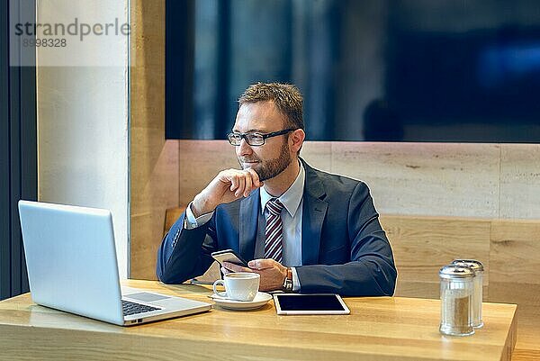 Geschäftsmann  der einen Kaffee genießt und mit einem Lächeln sein Handy auf Nachrichten überprüft  während sein Laptop und sein Tablet offen auf dem Tisch liegen