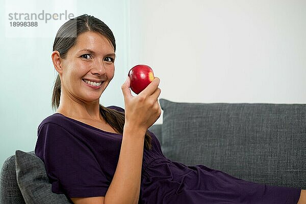 Hübsche junge Frau  die einen frischen knackigen roten Apfel in der Hand hält  während sie sich zu Hause auf dem Sofa entspannt und der Kamera ein freundliches Lächeln schenkt