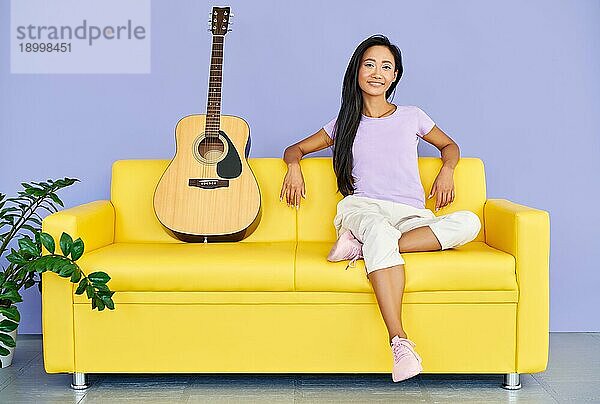 Hübsche Frau mit Gitarre sitzt auf einem bequemen Sofa und schaut in die Kamera